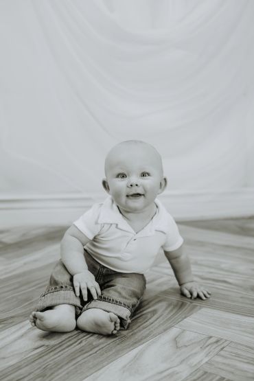 Smiling baby sitting on wooden floor in a black and white photo.