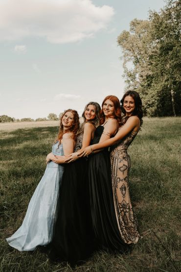 Four young women in elegant dresses smiling and posing together outdoors.