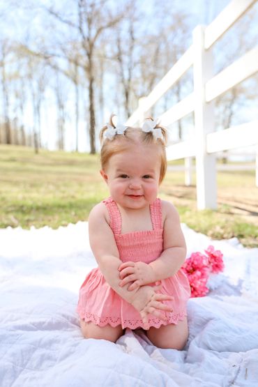 Smiling baby girl in a pink dress sitting on a blanket outdoors.