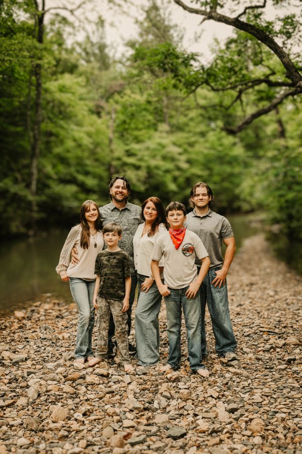 Smiling family of six standing barefoot on rocky ground near a lush green forest.