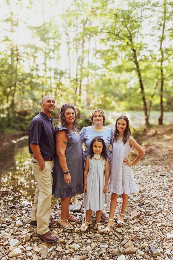 A family of five posing outdoors near a creek on a rocky ground.