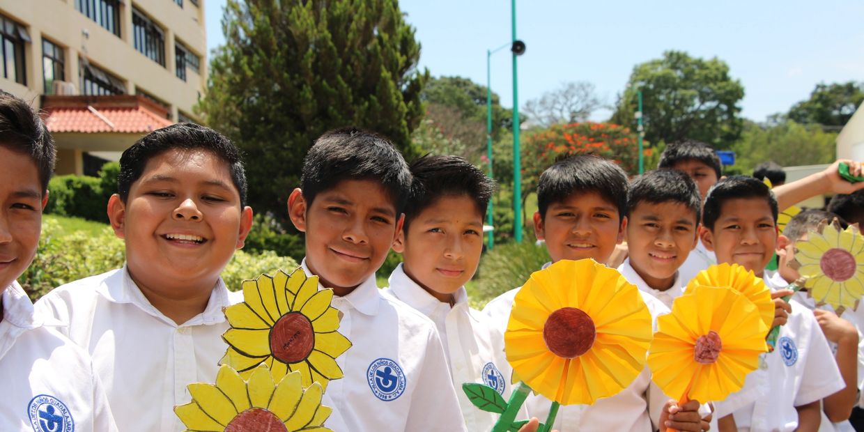 Smiling boys in white uniforms holding handmade paper sunflowers outdoors.