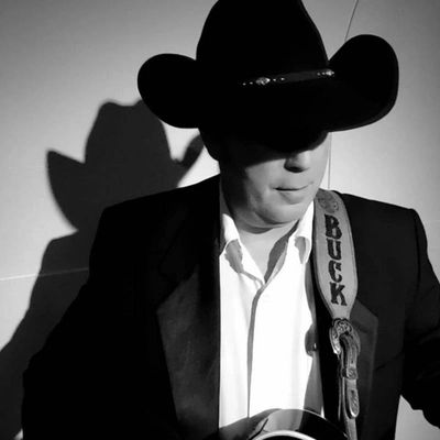 Man in cowboy hat playing guitar with 'BUCK' strap in black and white photo.