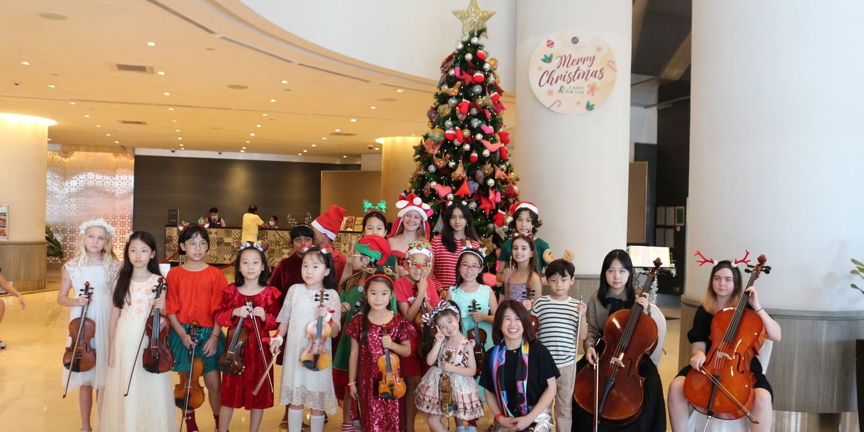 Children musicians in festive attire posing with instruments by a decorated Christmas tree.
