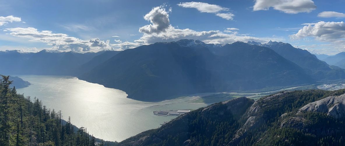 Panoramic view of a sunlit lake surrounded by forested mountains under a partly cloudy sky.