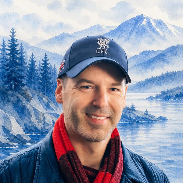 Man smiling in front of a scenic mountain and lake background, wearing a Liverpool FC cap and red scarf.
