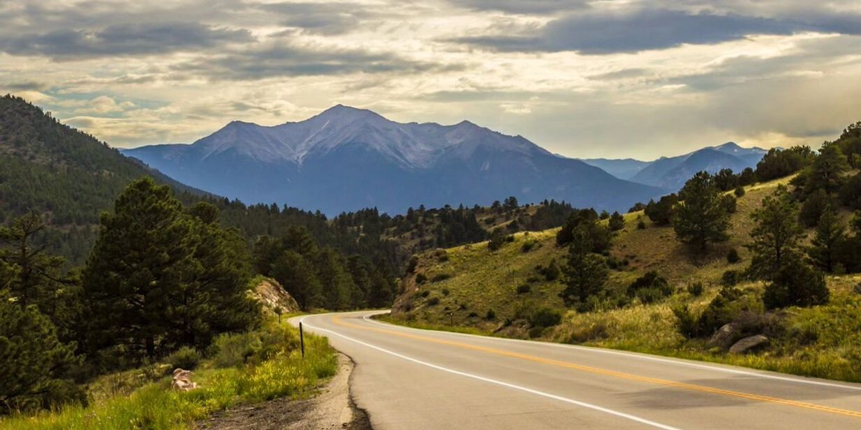 Winding rural road lined with dense forest leading toward a snow-covered mountain in the distance.
