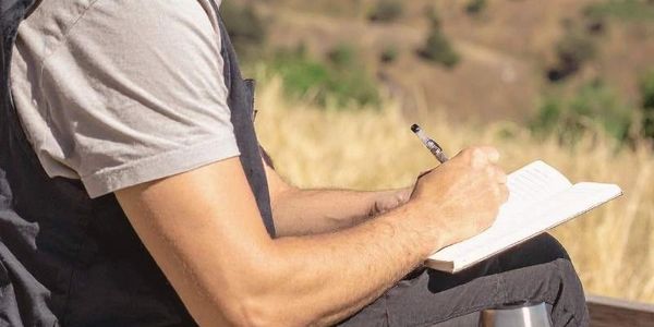 Man writing in a journal outdoors with mountains in the background during a reflective moment.