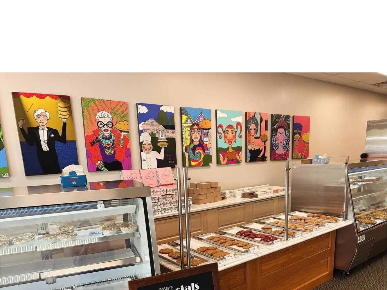 Colorful pie-themed artwork adorns the walls of a bakery with cookies and pies on display.