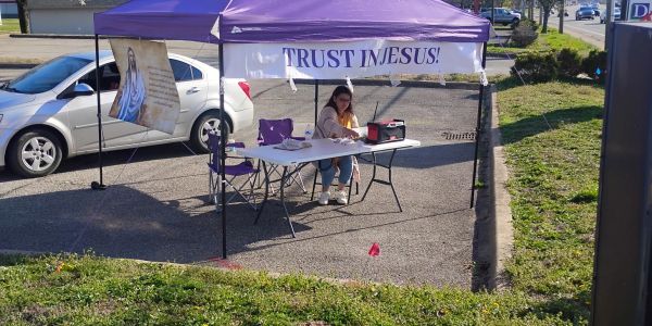 A woman sits under a purple tent with a 'Trust in Jesus!' banner in a parking lot.