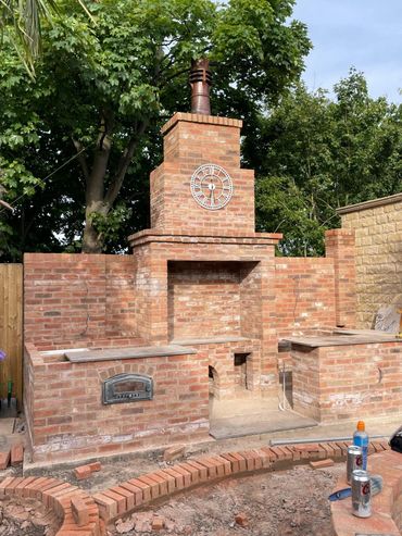Outdoor brick fireplace with clock and pizza oven under construction.