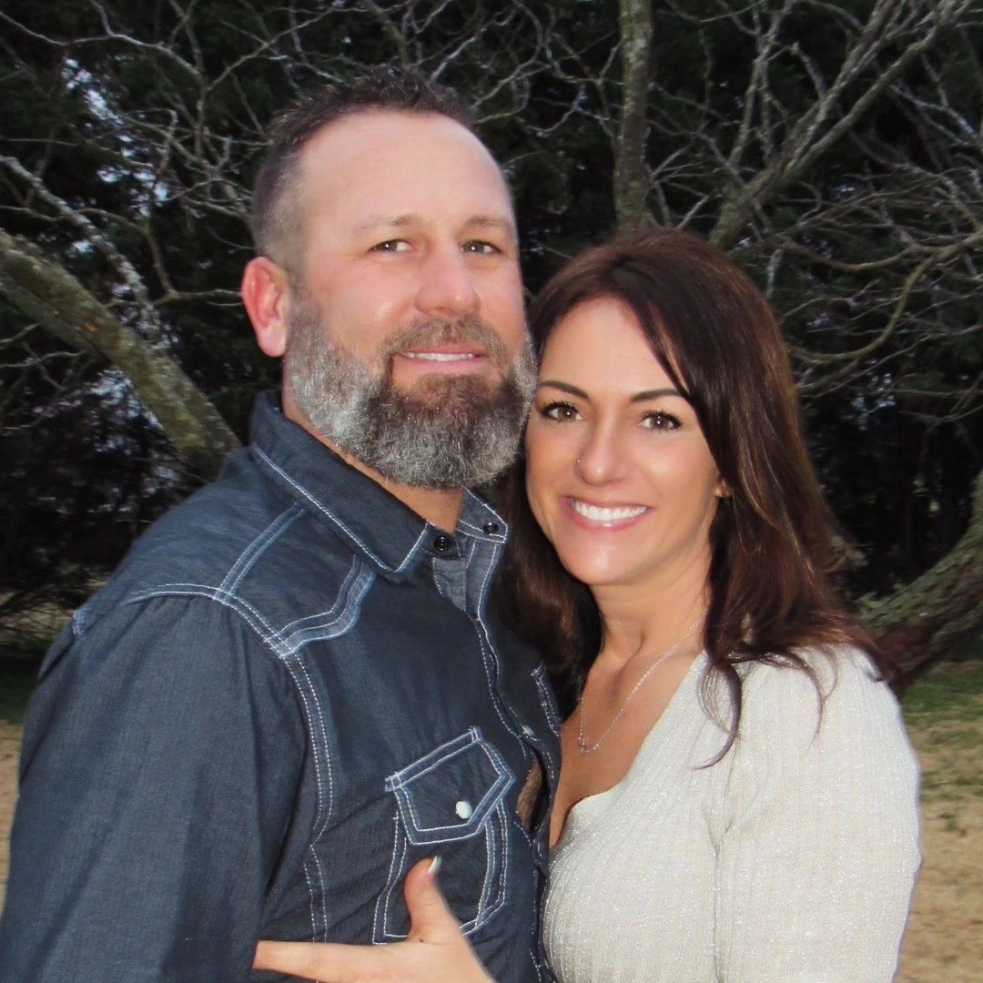 A smiling couple posing outdoors in front of leafless trees.