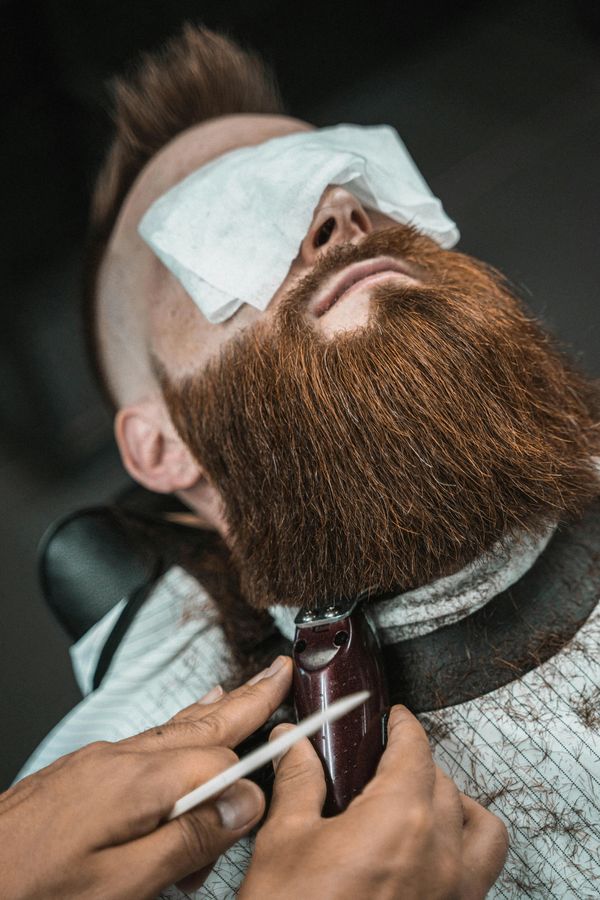 Man getting his thick red beard trimmed with electric clippers.