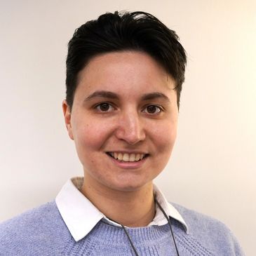 Smiling person with short dark hair wearing a light blue sweater and white collared shirt.