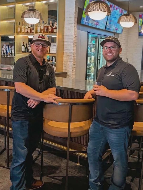 Two men in casual attire stand and smile in a modern bar setting.
