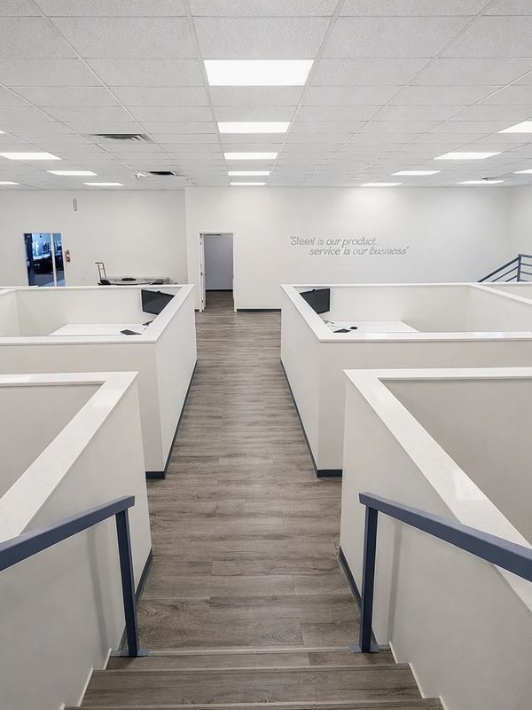 Modern office cubicles with white walls and wooden flooring under bright ceiling lights.