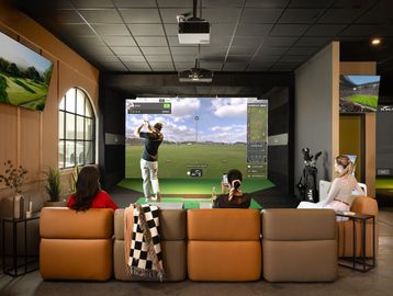 Four people watch a golf simulator in a cozy room with modern decor.
