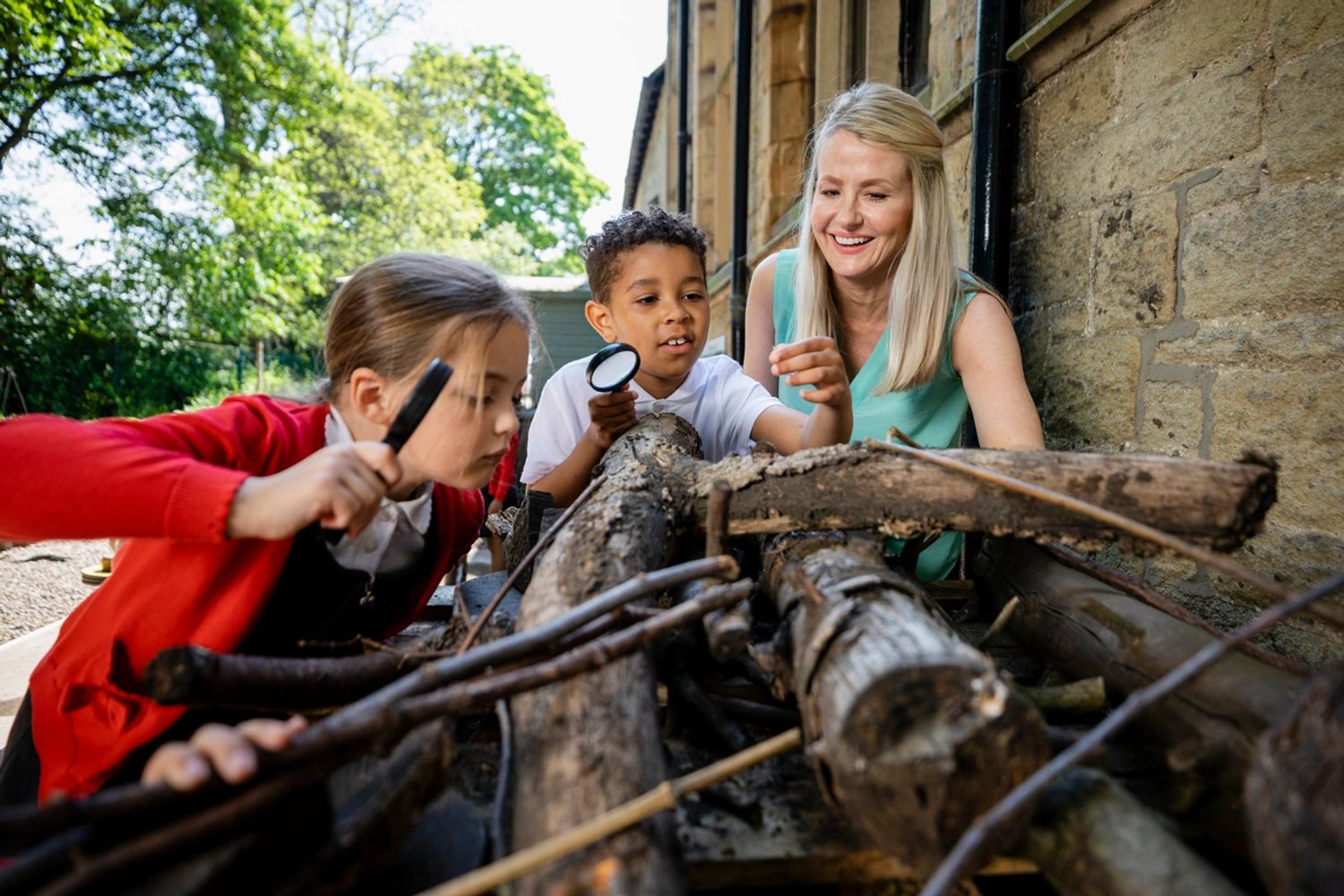 Children and teacher explore nature with magnifying glasses outdoors.