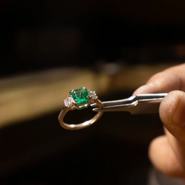A hand holding tweezers with an emerald and diamond ring.