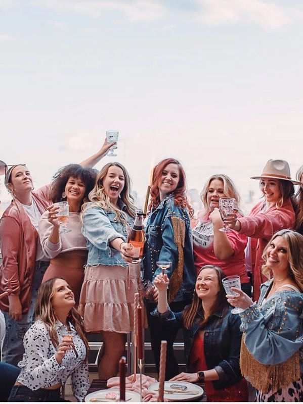 Group of women in pink and blue jean attire making a toast with champagne on a rooftop
