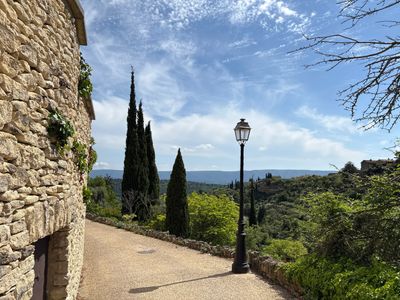 A peaceful stone pathway with tall cypress trees and a vintage street lamp under a partly cloudy sky.