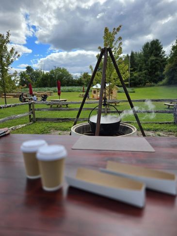 Outdoor cooking pot hanging over fire pit on a wooden table with coffee cups.