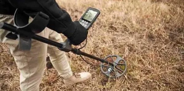 Person using a metal detector in dry grass.