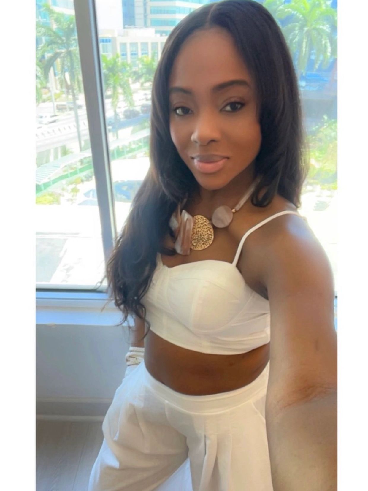 Woman in white outfit takes selfie indoors with cityscape background.