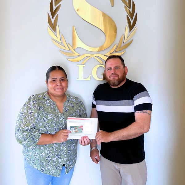 Two people holding a certificate in front of a gold emblem on the wall.