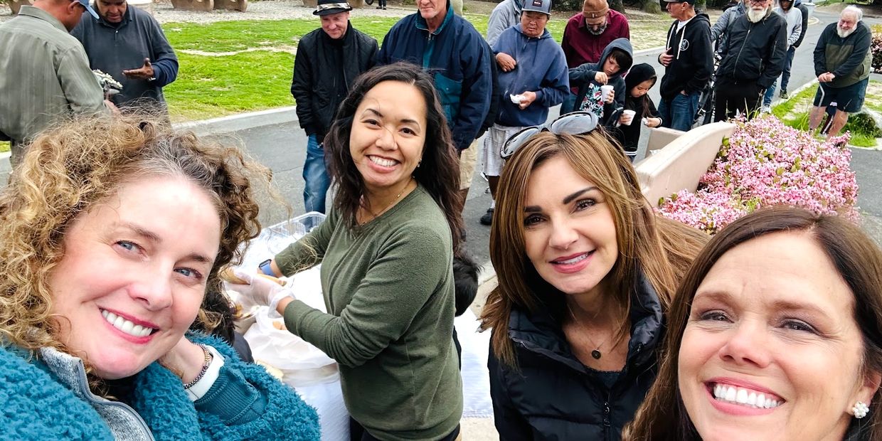 Four women smiling at a community event with people lined up in the background.