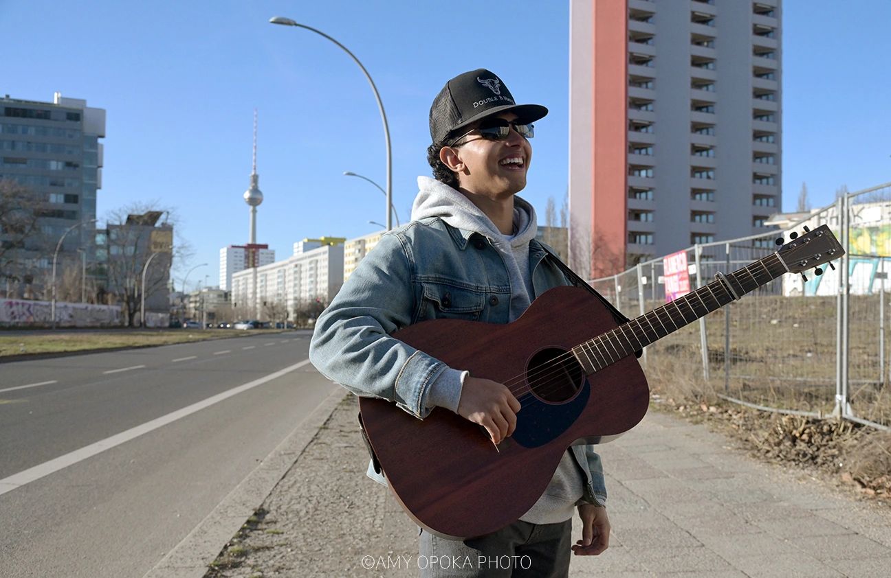 Young man playing guitar and smiling on a city sidewalk under clear blue sky.