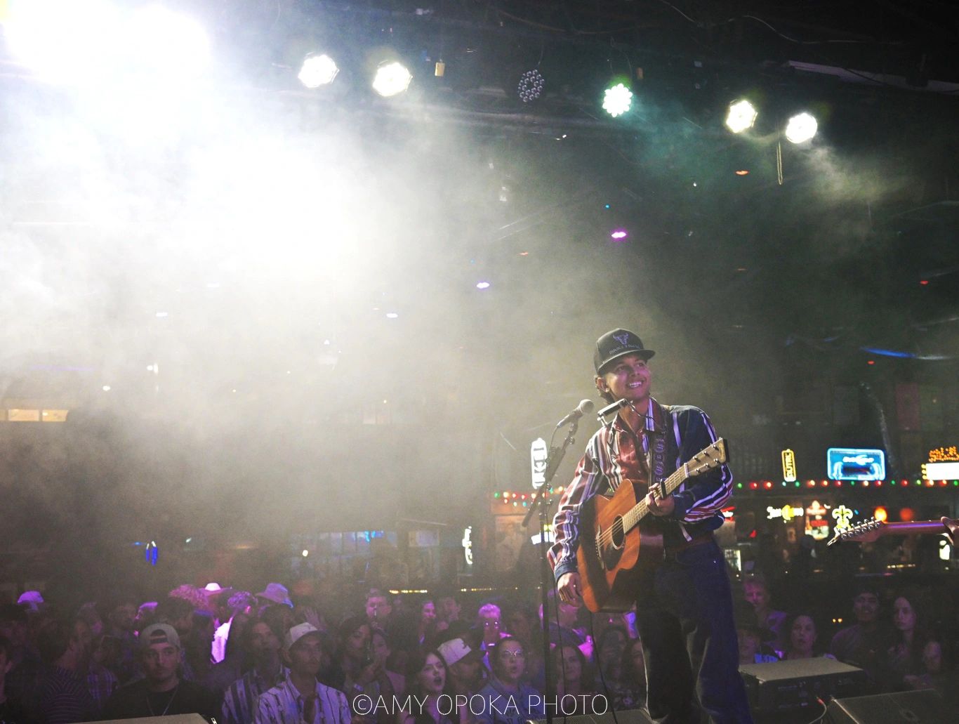 Musician plays guitar on stage amid bright lights and fog in front of a crowd.