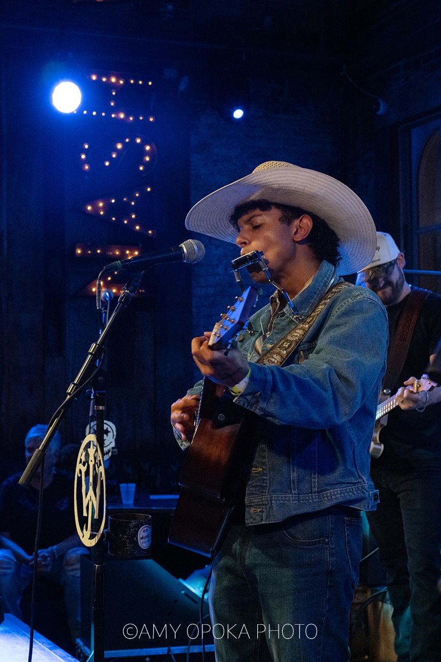 Musician in a cowboy hat playing guitar and harmonica on stage.