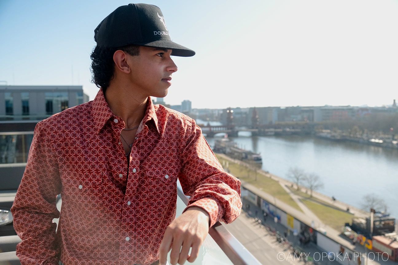 Man in a patterned shirt and cap overlooking a river from a balcony.