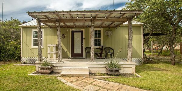 A rustic green cabin with a covered porch, and cozy outdoor seating viewing a natural landscape.