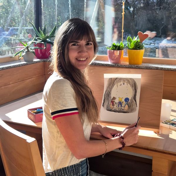 A woman smiling while painting a colorful illustration at a wooden desk near a sunny window.