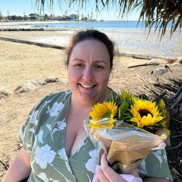 A smiling woman holding sunflowers at a sunny beachside location.