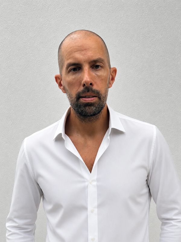 Man with a shaved head and beard wearing a white dress shirt against a gray wall.