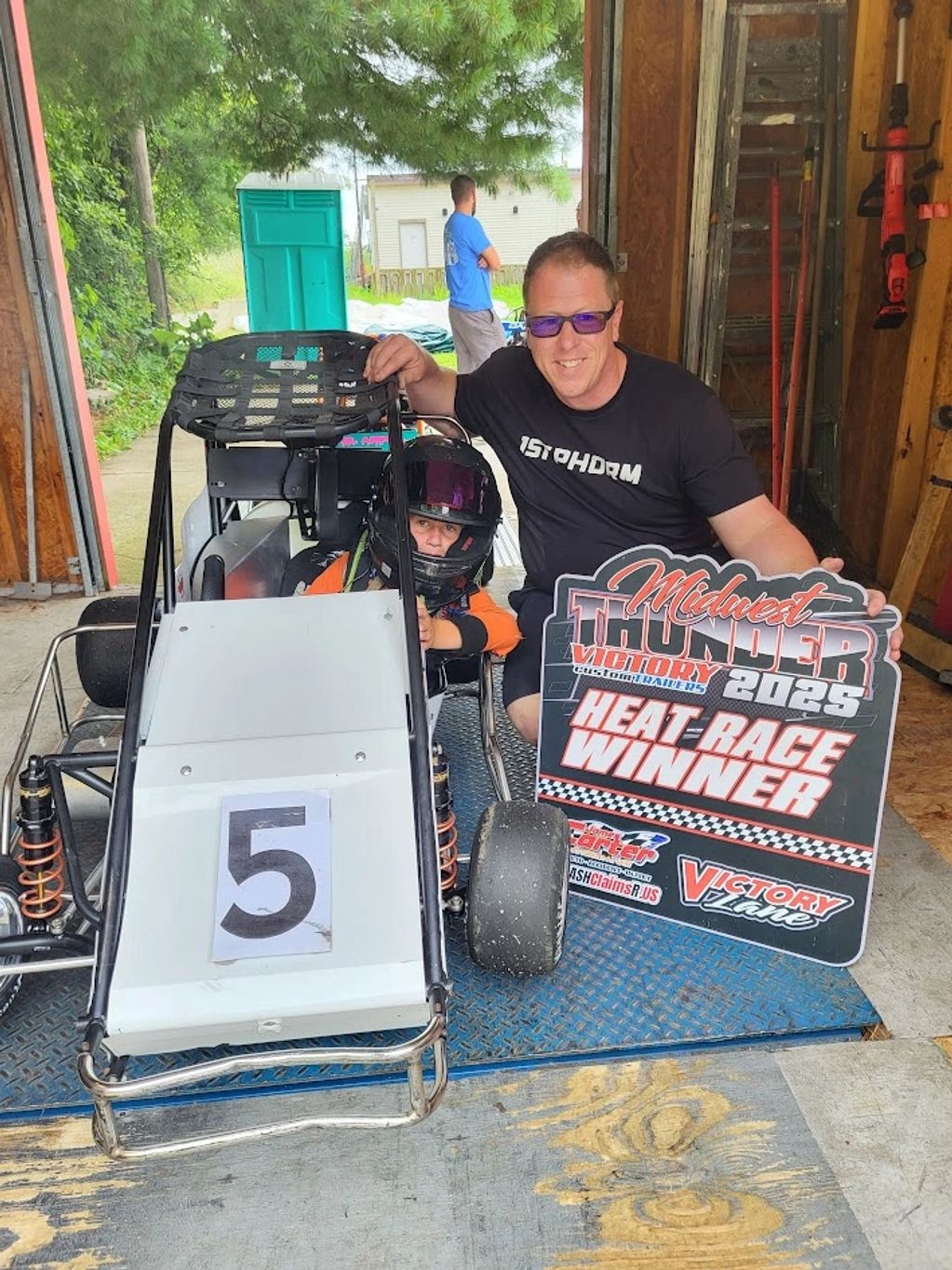 Young racer in a go-kart with an adult holding a 'Heat Race Winner' sign.