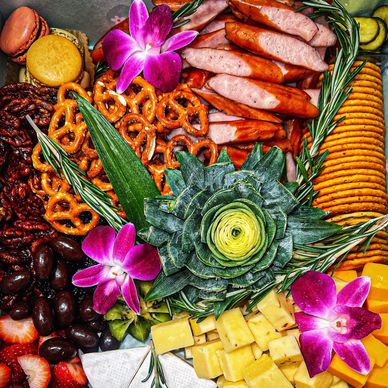 Colorful charcuterie board with pretzels, cheese, sausages, and flowers.