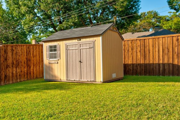 A small beige garden shed with double doors and a window in a grassy yard.