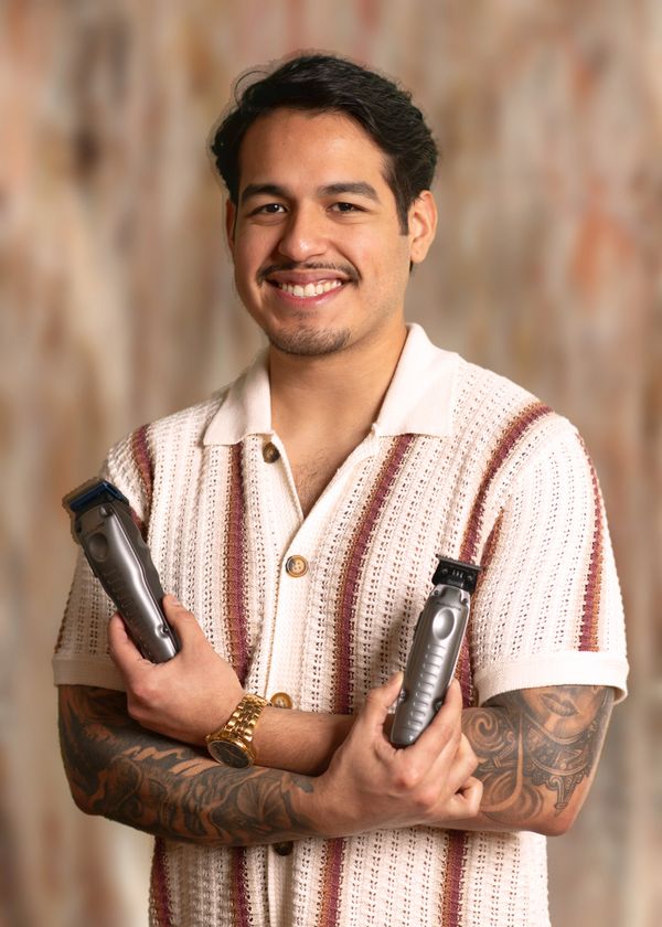 Smiling man holding two hair clippers, showing off tattoos and wearing a striped shirt.