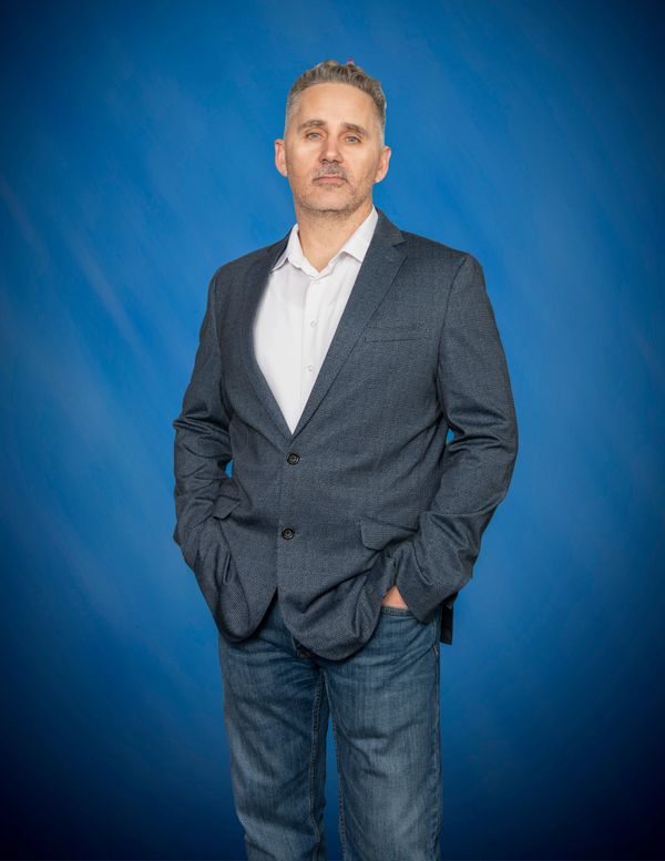 Man in gray blazer and jeans against blue background.