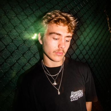 Young man with blonde hair and mustache wearing layered necklaces and a black T-shirt.