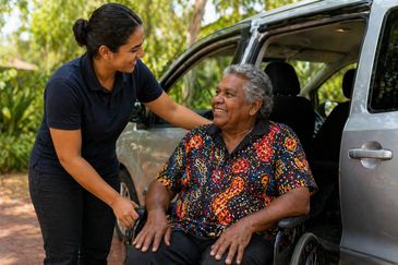 Young woman helping elderly woman in wheelchair by a car.