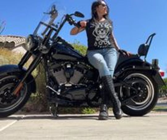 Woman posing confidently next to a black motorcycle under clear blue sky.