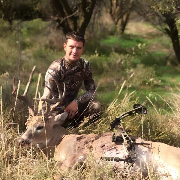 Hunter posing with a bow and a deer in a grassy field.