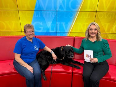 Two women and a black dog sitting on a red couch in a TV studio.