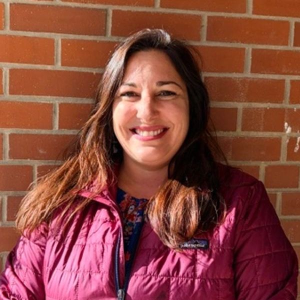 Woman smiling in a maroon jacket against a brick wall.