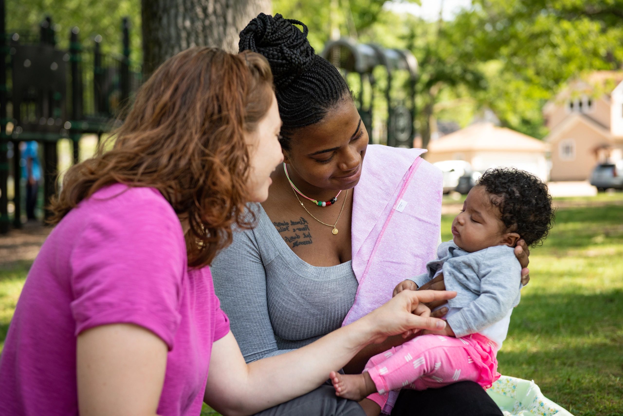 Two women engaging with a baby outdoors on a sunny day.